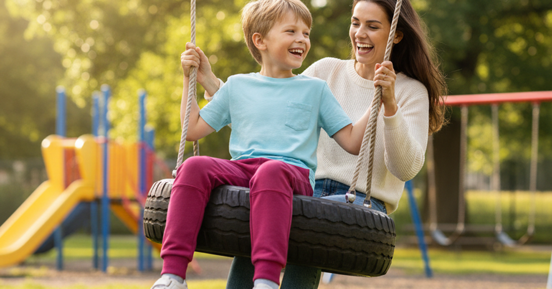 Active boy in double-knee joggers on a tire swing at the playground with his mom